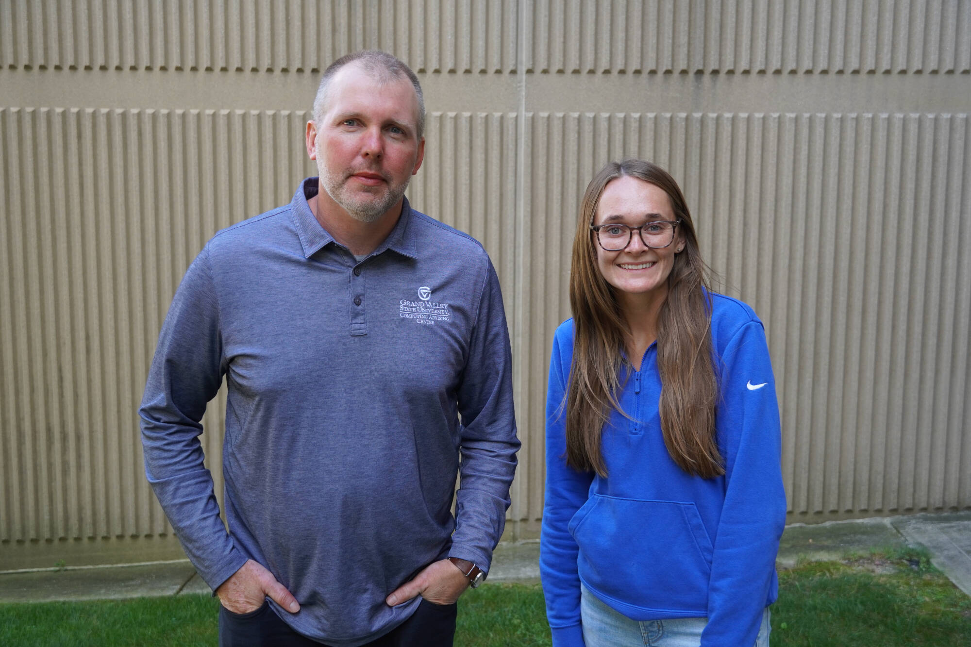 Colin DeKuiper and Emily Zeliasko, academic advisors in the GVSU College of Computing, standing outside against a tan textured wall. Colin is wearing a Grand Valley State University polo shirt, and Emily is wearing a blue pullover and glasses, smiling.